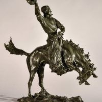 Alt text: Bronze sculpture of a rearing bronco with a cowboy on his back, holding his hat off his head with his right hand