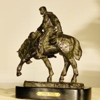 Alt text: Bronze sculpture of a cowboy riding on a horse with its front left leg outstretched and head down, angled view