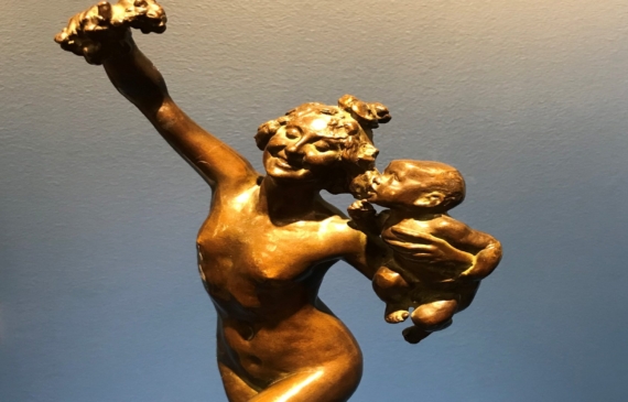 Alt text: Bronze sculpture of a woman holding fruit in the air in one hand, and a baby in the other