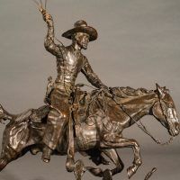 Alt text: Bronze sculpture of a cowboy with a lasso riding a horse jumping over a cactus, detail side view