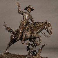 Alt text: Bronze sculpture of a cowboy with a lasso riding a horse jumping over a cactus, angled view