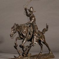 Alt text: Bronze sculpture of a cowboy with a lasso riding a horse jumping over a cactus, angled view