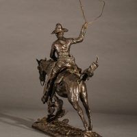 Alt text: Bronze sculpture of a cowboy with a lasso riding a horse jumping over a cactus, angled view