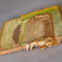 Alt text: Abstract paint and gilt patterns on burlap mounted to a wood board, angled view