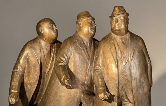 Alt text: Bronze sculpture of three businessmen in trench coats walking together, frontal view